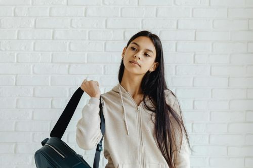 Teen girl in hoodie with backpack standing by a white brick wall, looking thoughtful.