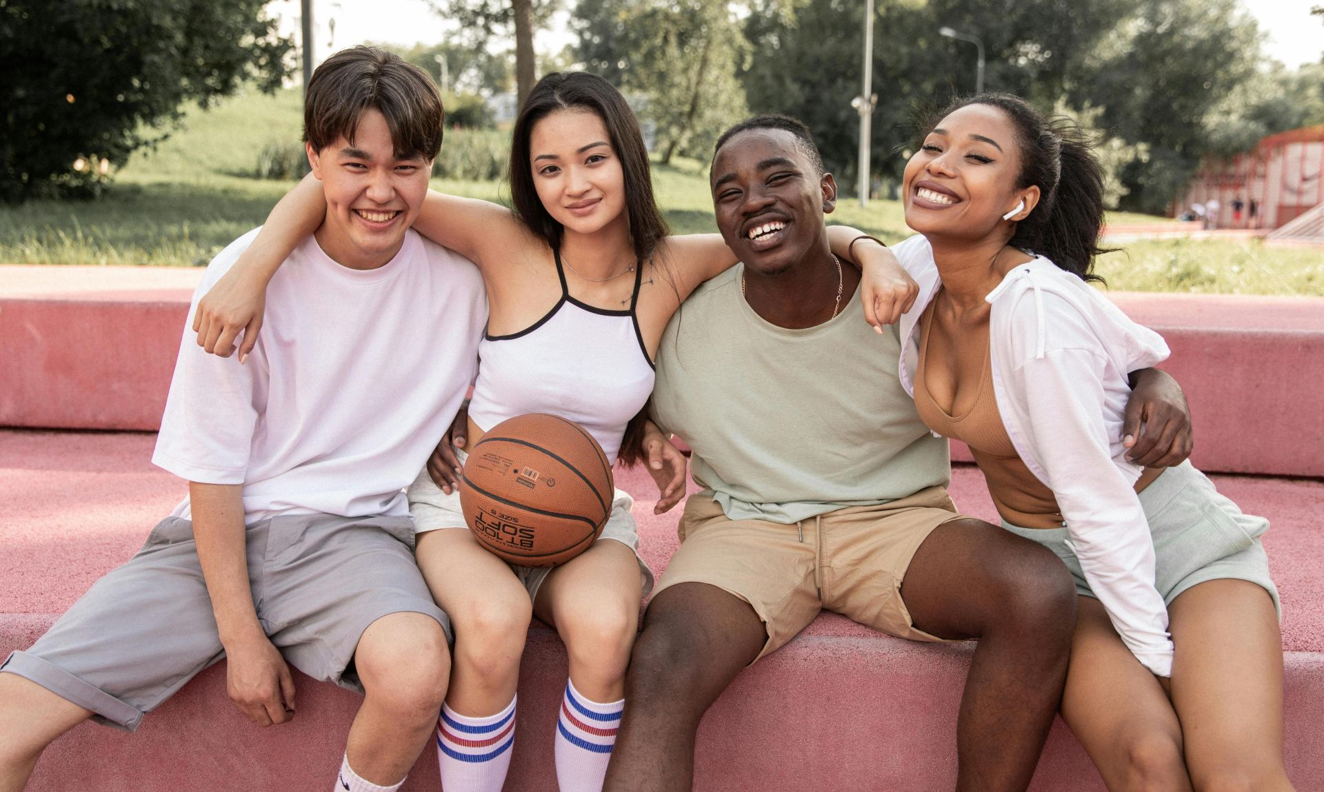 Content young multiracial friends wearing casual clothes embracing and looking at camera with happy smiles while sitting on bench in green park